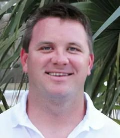 Man wearing a white collared shirt smiles. Palm fronds are behind him.