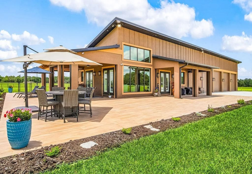 A modern, tan-colored house with a patio, outdoor dining set, and lush green lawn on a sunny day.