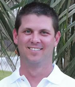 Man smiling, wearing a white collared shirt, against a blurred outdoor background.