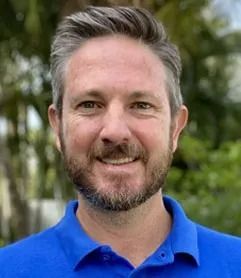 Man in blue shirt smiles, graying hair, outdoor setting.