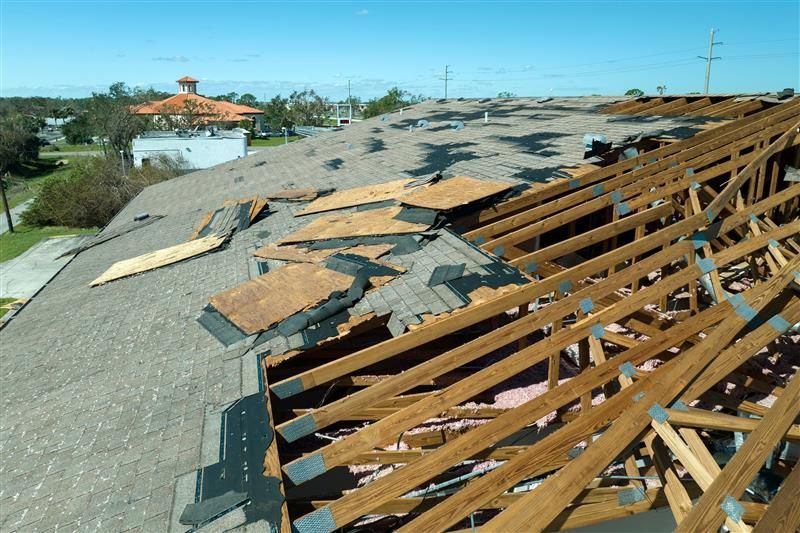 Damaged roof with exposed wooden beams, remnants of shingles, and a sunny, clear sky in the background.