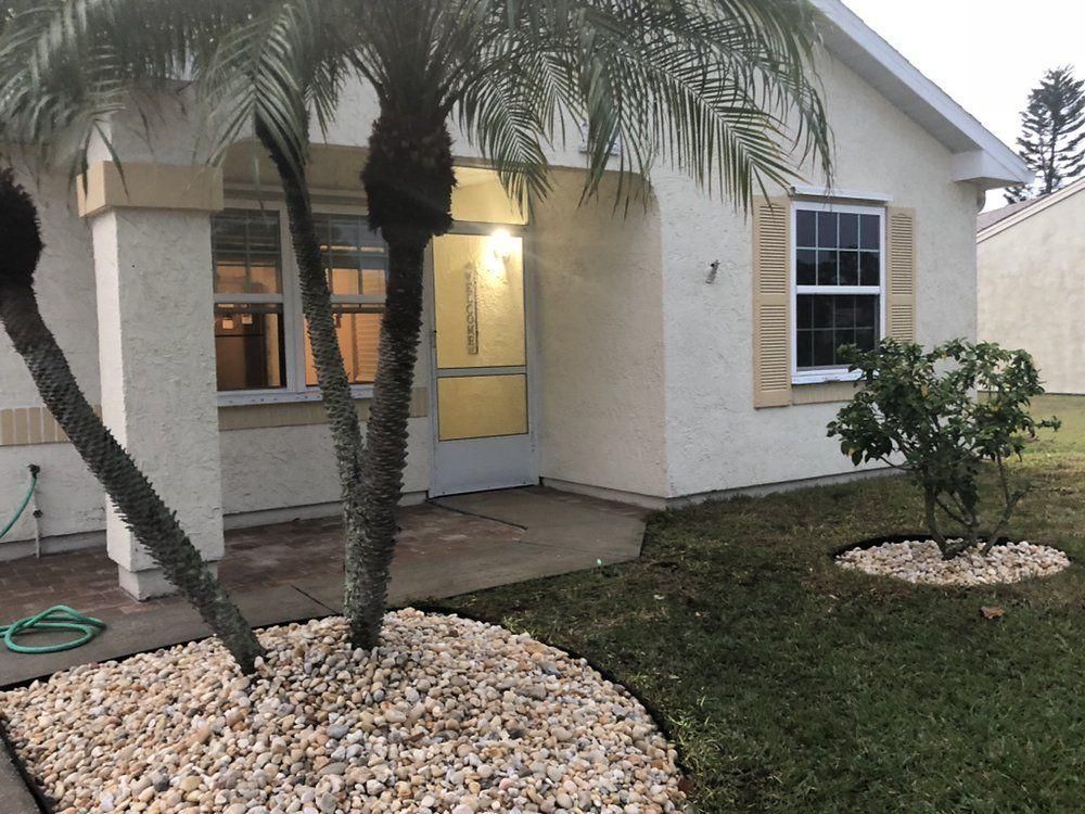 Front of a beige house with a yellow door, windows, palm trees, and rock landscaping