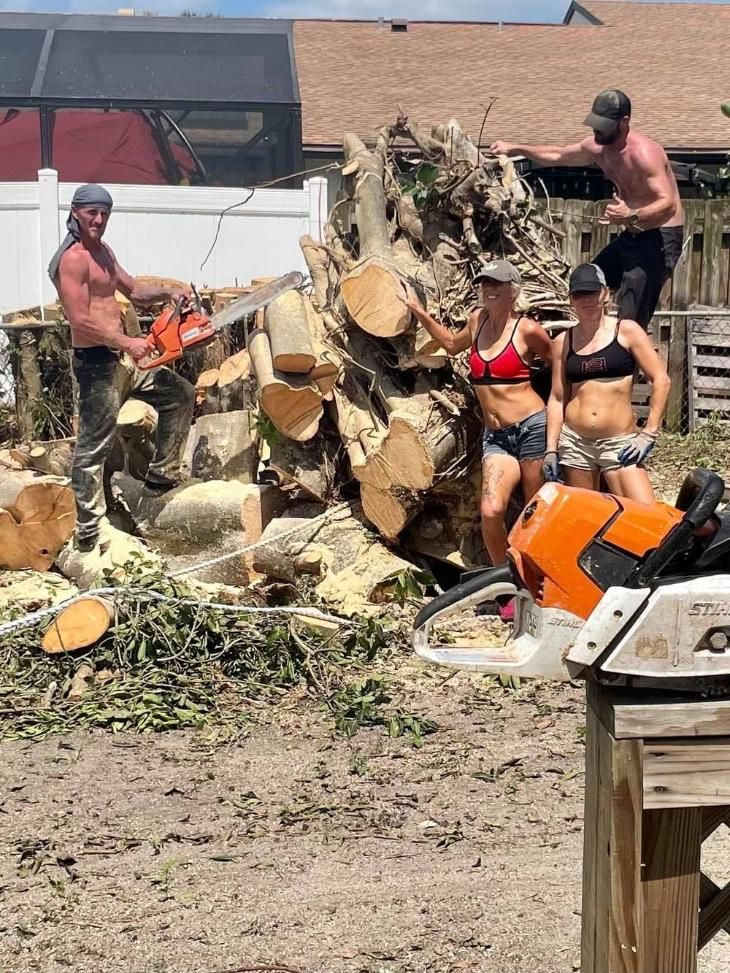 Workers clearing a fallen tree trunk in a muddy yard with a chainsaw and wood chips scattered around.
