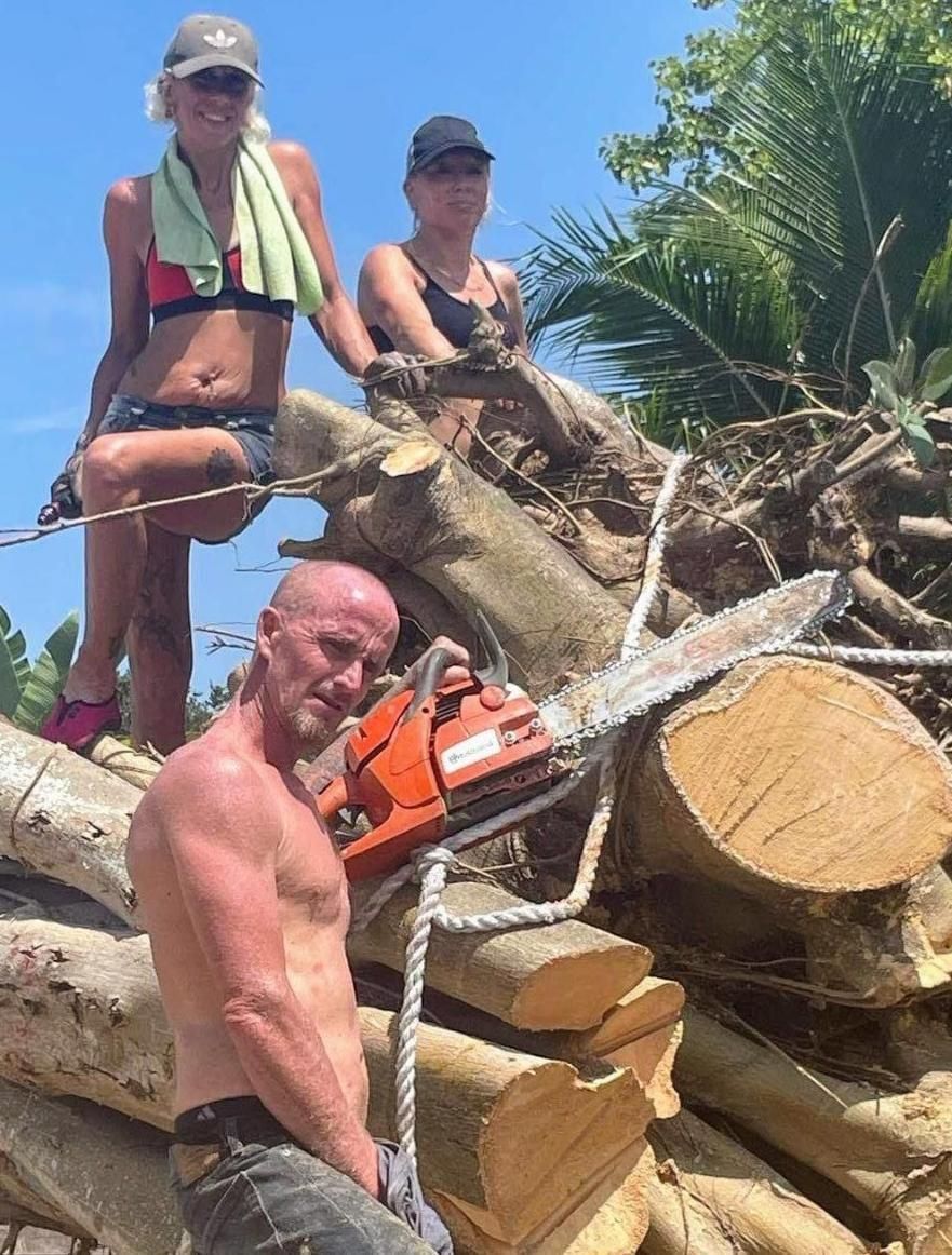 Three workers posing on a large pile of cut logs beside a chainsaw in a sunny tropical setting