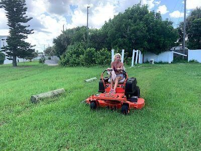 Person riding an orange lawn mower on a grassy field near trees and a white building.