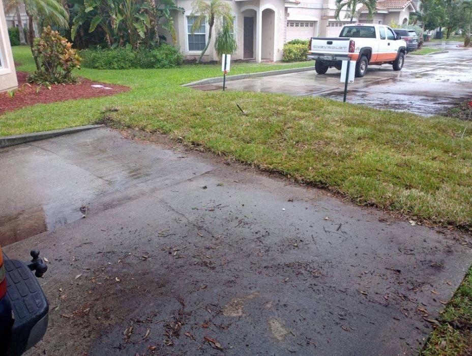 Rainy driveway and front yard with a wet sidewalk, grass, and a pickup truck parked by a house.