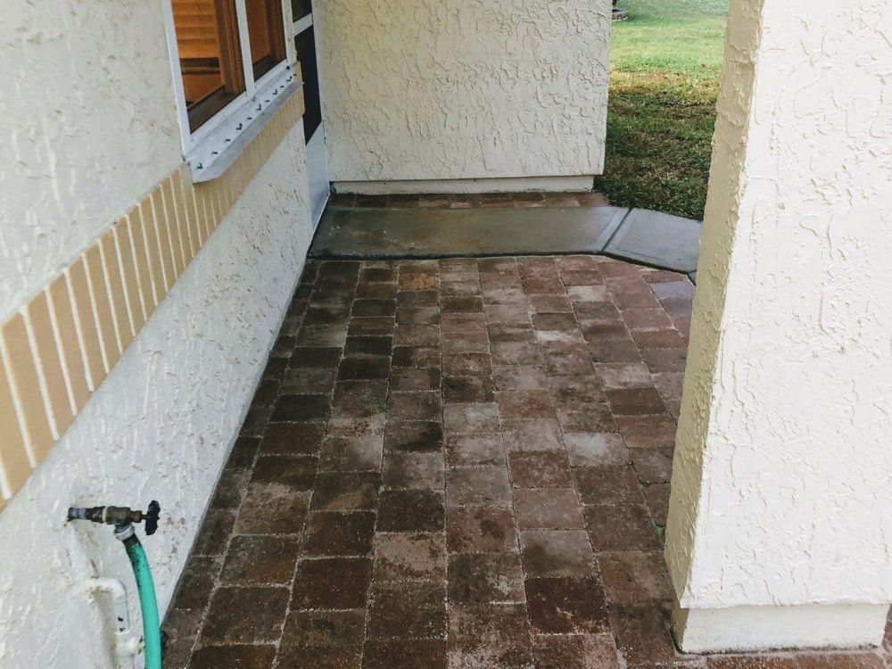 Narrow outdoor tiled patio beside a stucco house, with a hose spigot and grass at the end.