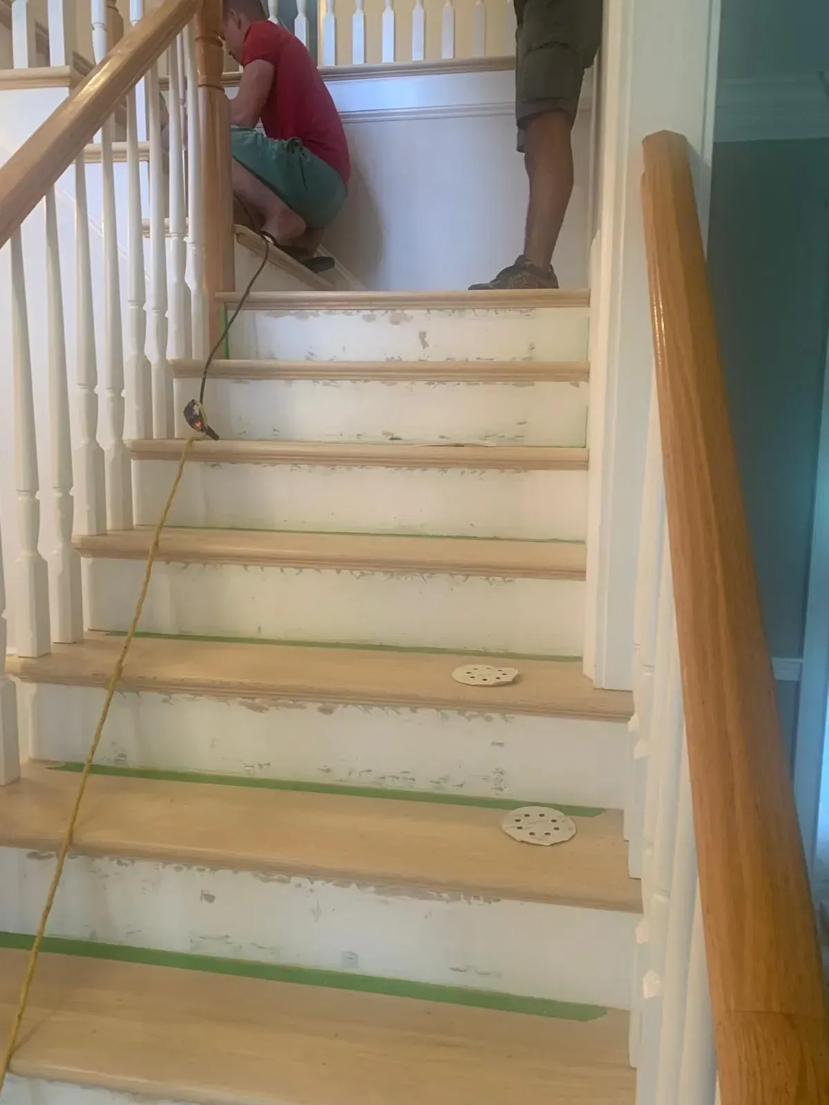 People working on a wooden staircase; one kneeling, one standing; steps are unfinished, white and green tape.