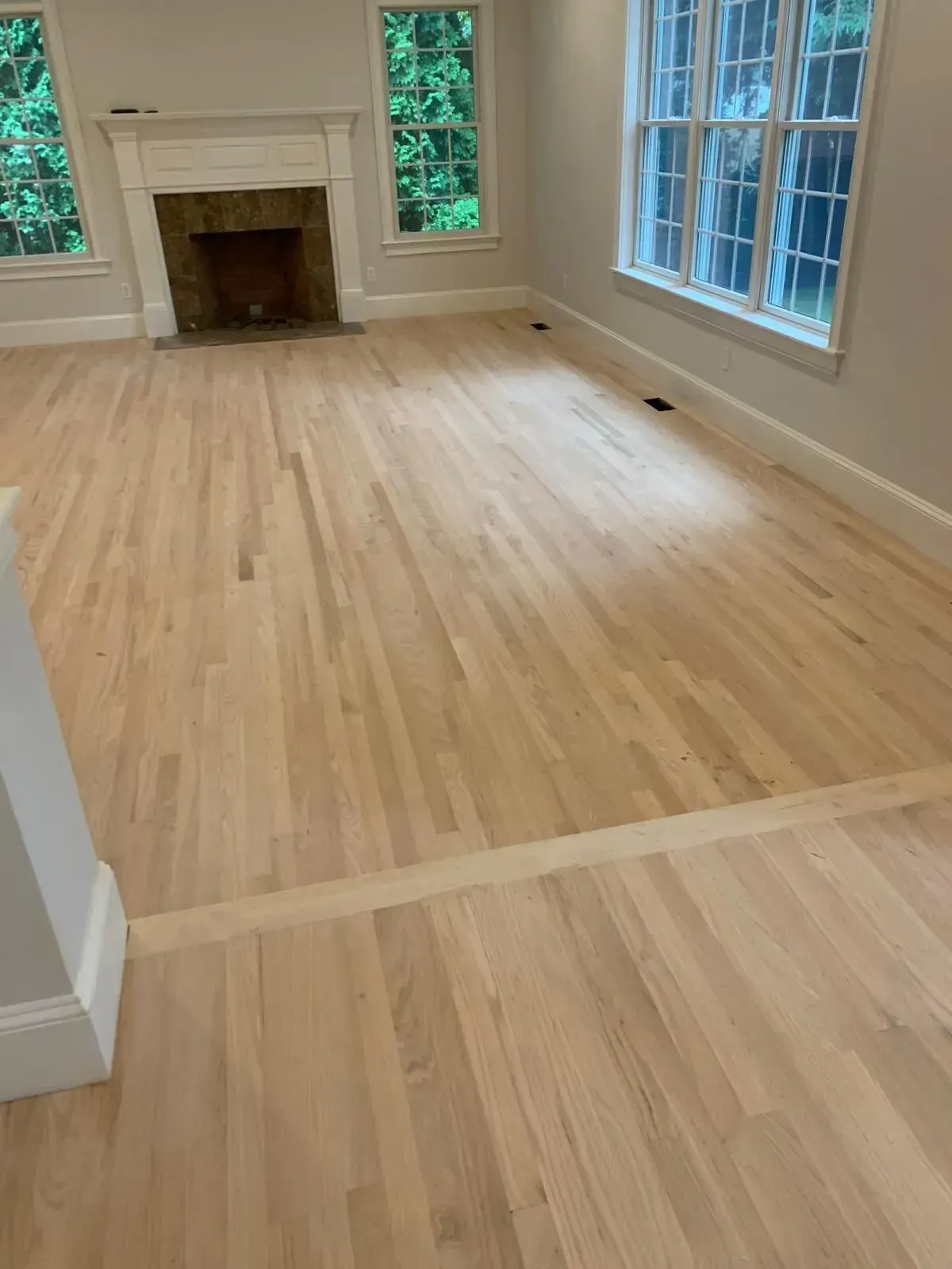 Light-toned hardwood floors in a sunlit living room with a fireplace and windows.