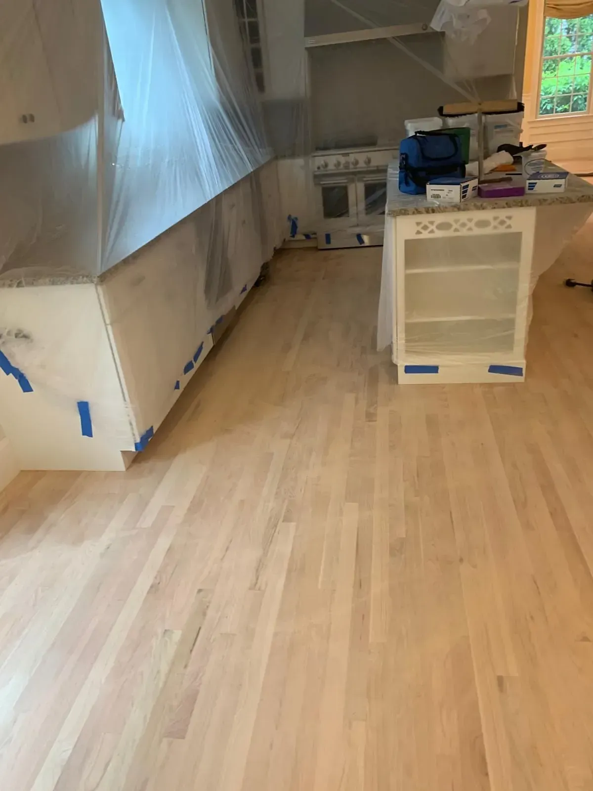 Light-colored wood floor in a kitchen under renovation; cabinets and island covered in plastic, oven visible.