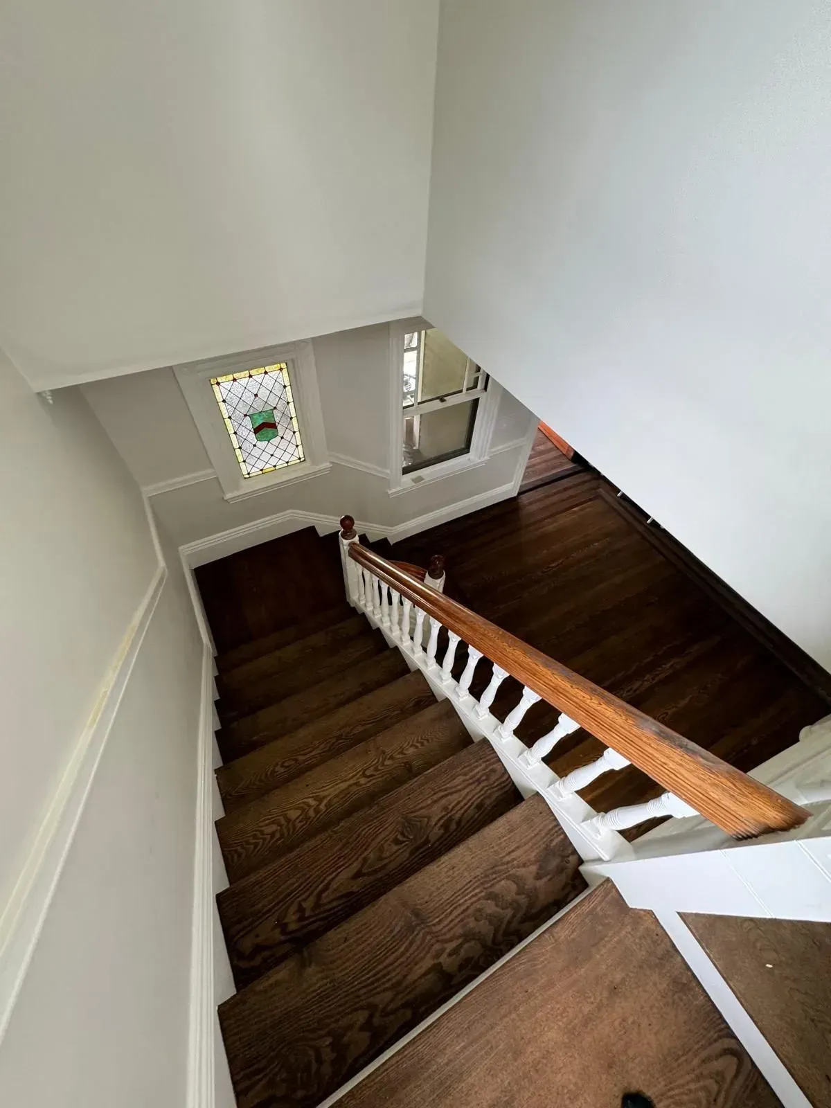 View down a wooden staircase with a white banister, stained glass window, and light gray walls.