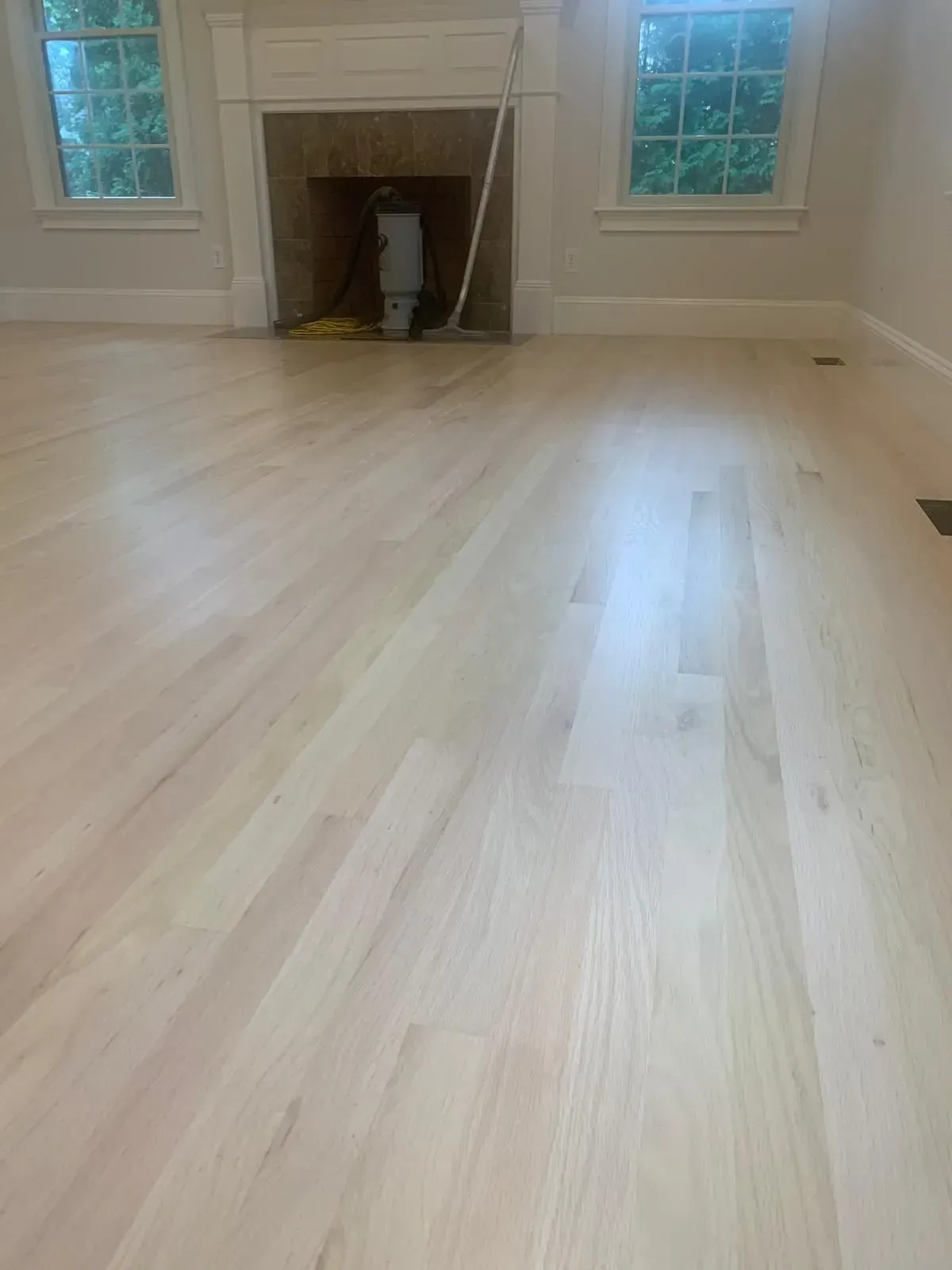 Light wood floors in a room with a fireplace and two windows.