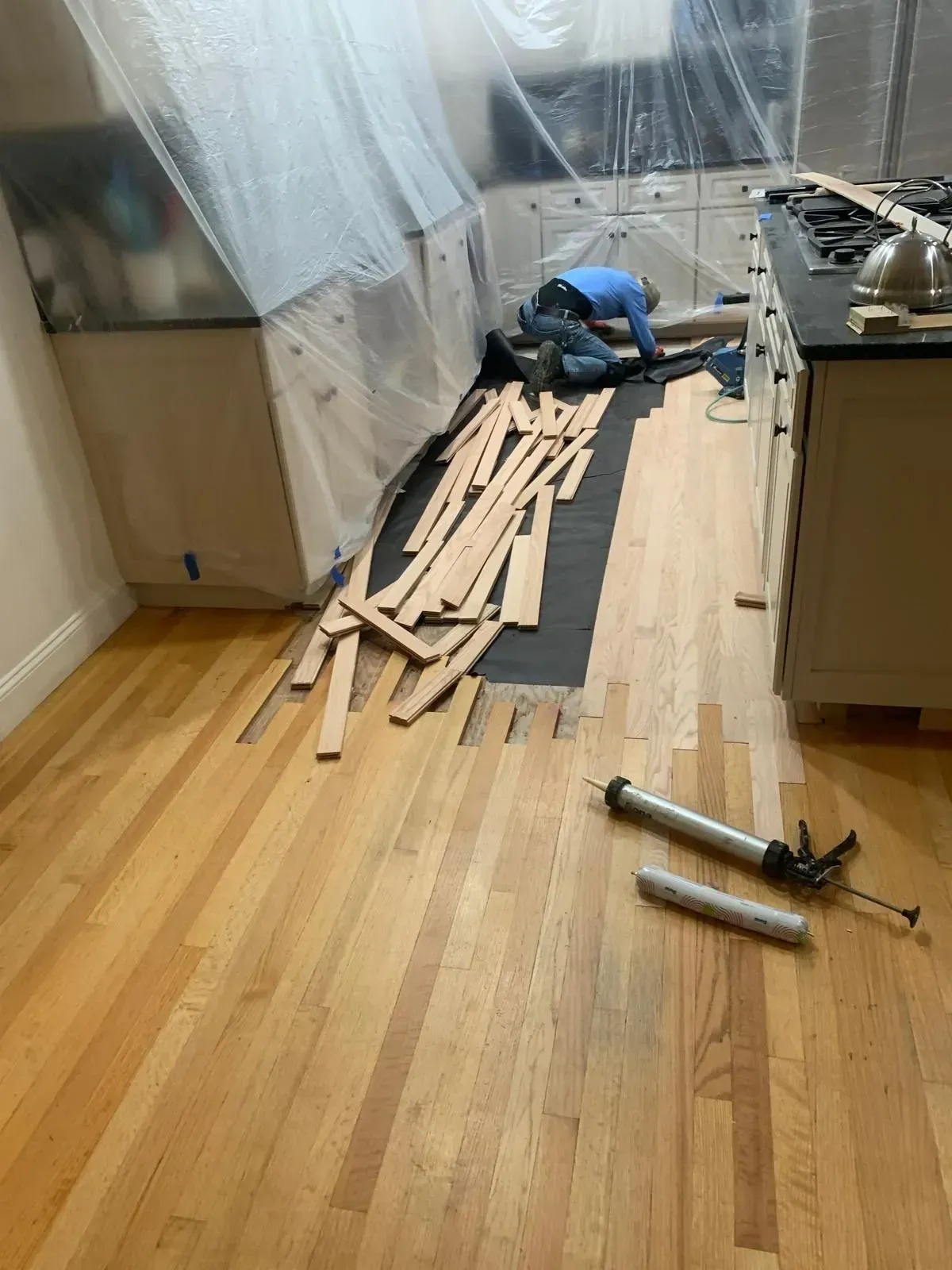 Kitchen floor renovation: worker installs wood planks. Plastic sheeting protects cabinets. Tools and loose planks on floor.