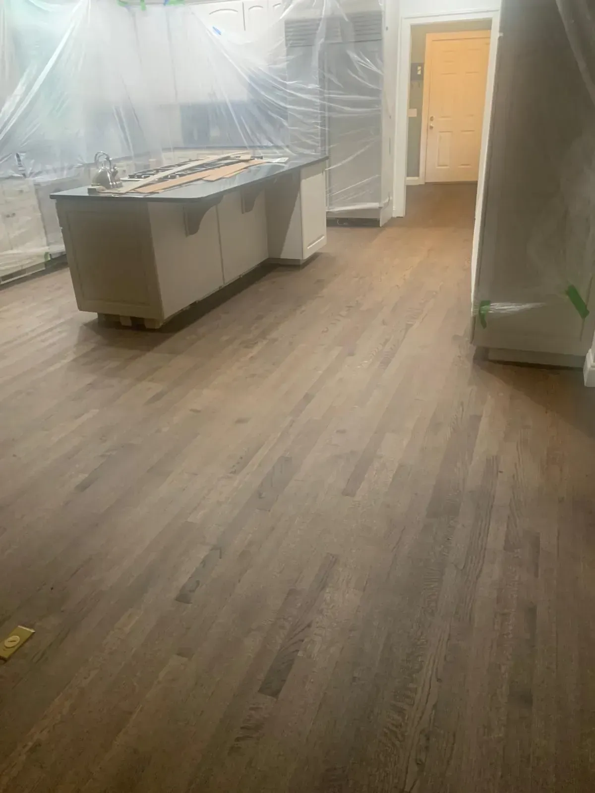 Wooden floor in a kitchen, stained a brownish color. The island and cabinets are covered in plastic.