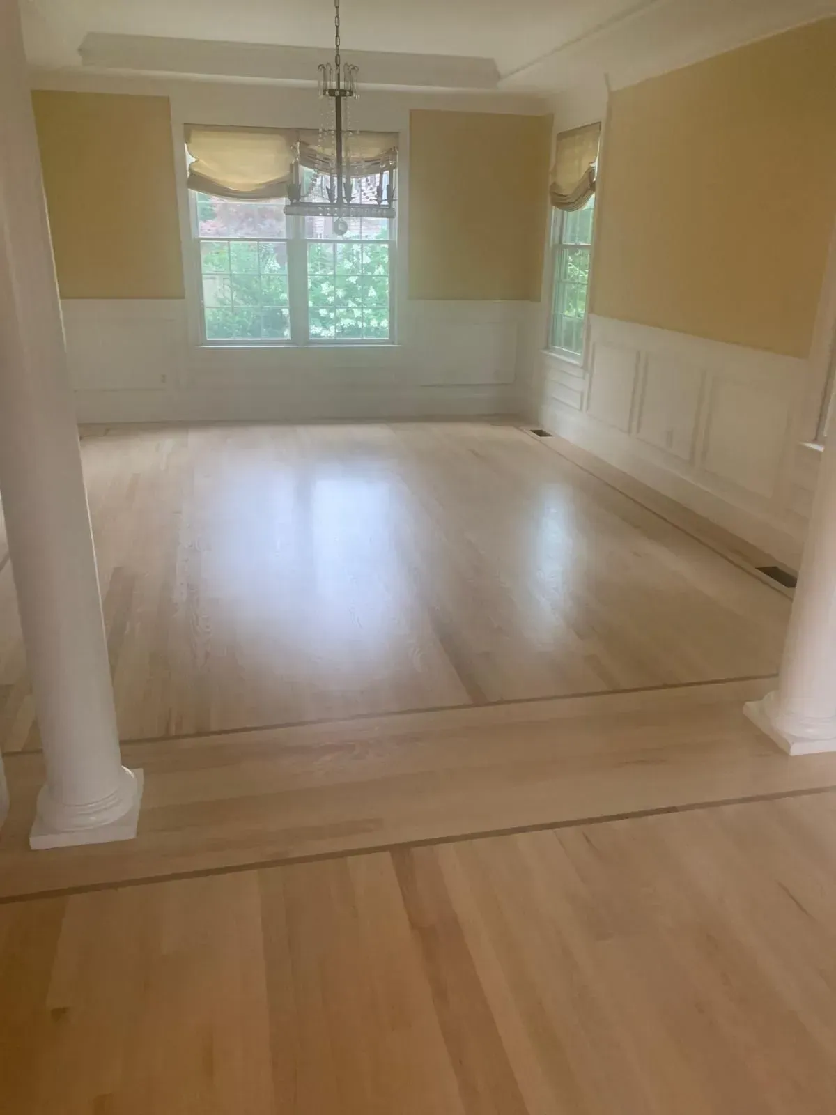 A light-filled room with light wood floors, yellow walls, and large windows; two white pillars in foreground.