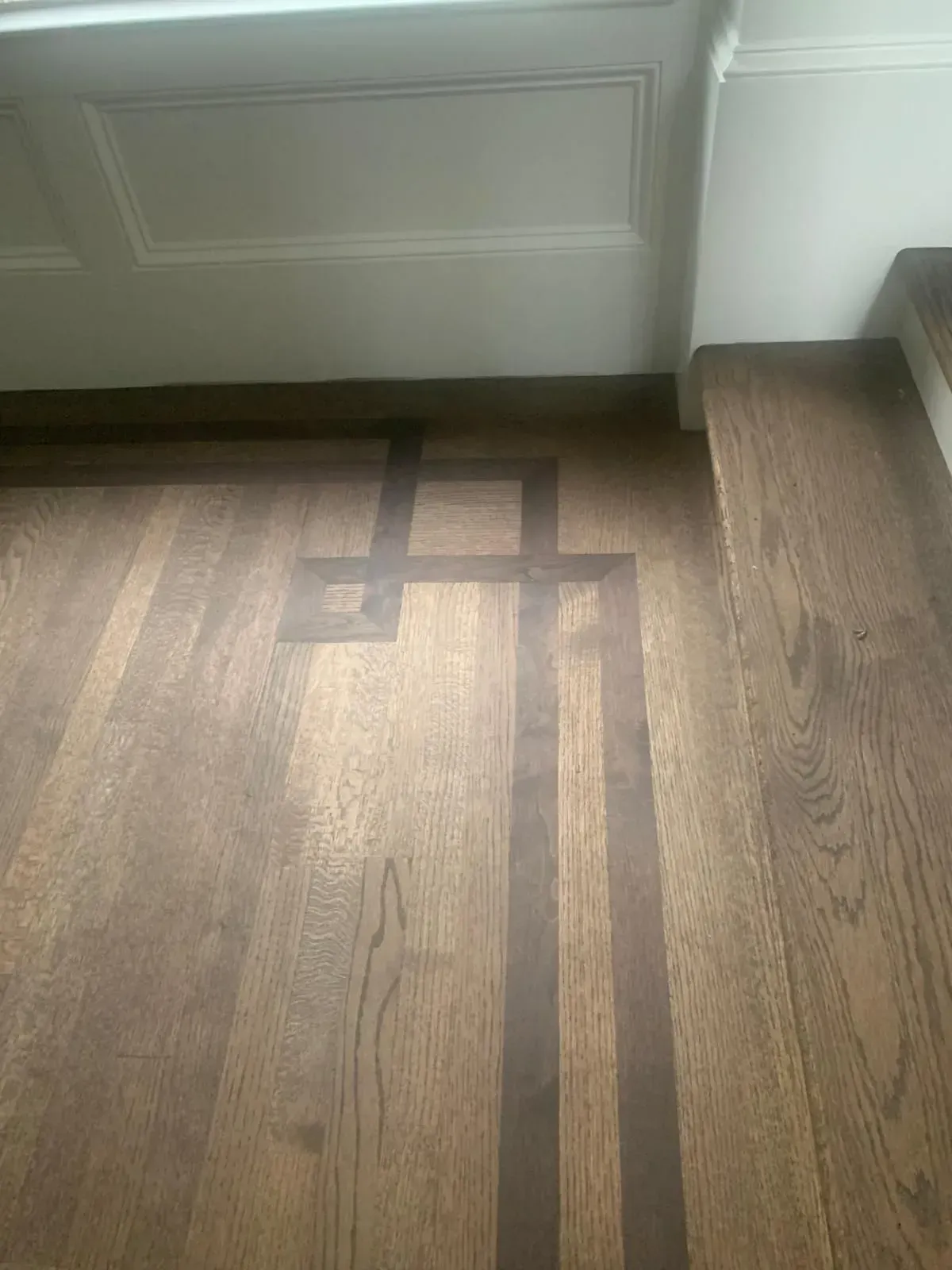 Brown hardwood floor with dark brown decorative inlay pattern, adjacent to white wall trim.