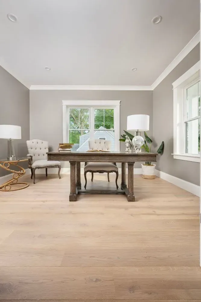 A home office with a wooden desk and chairs, light gray walls, and blonde wood floors.