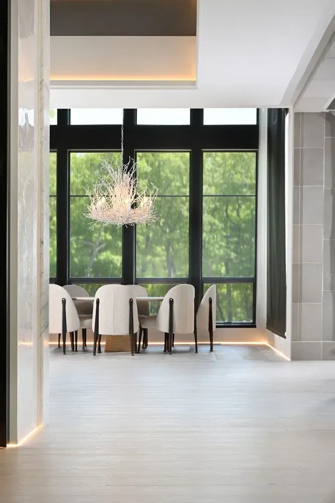 Dining room with table and chairs, chandelier, large window, green trees outside.