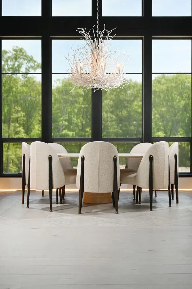 Dining room with a table, chairs, and a chandelier in front of large windows overlooking a green forest.