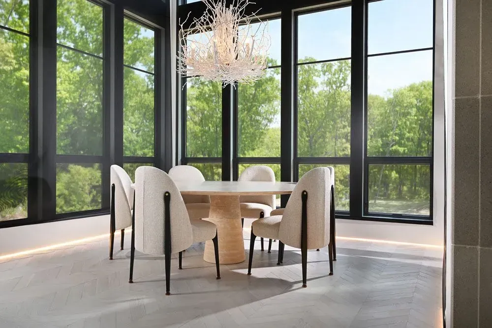 Dining room with table, chairs, and large windows overlooking trees. Chandelier hangs above.