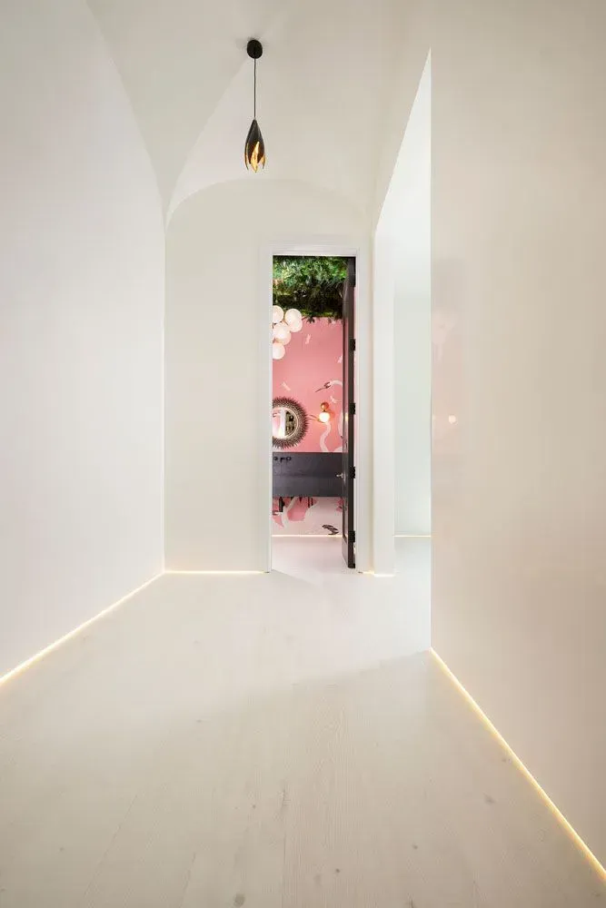 White hallway with wood floors, leading to a pink room with decor; single hanging light, light strip along floor.