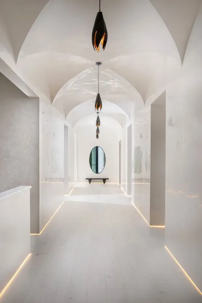 Long, white hallway with arched ceiling, lined with lights, and an oval mirror at the end.