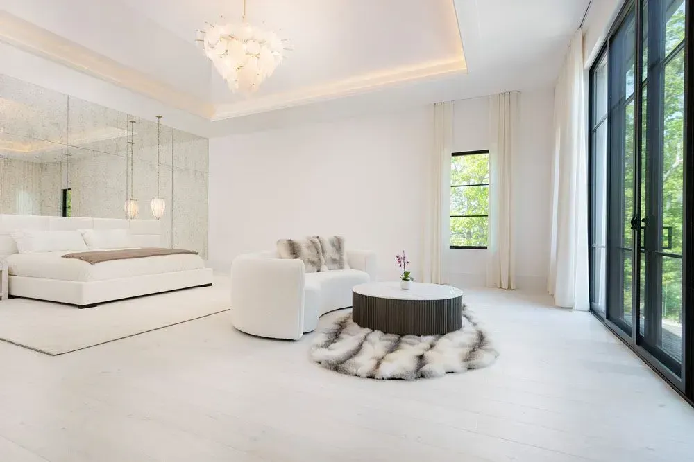 White bedroom with a bed, seating area, large windows, and a chandelier.