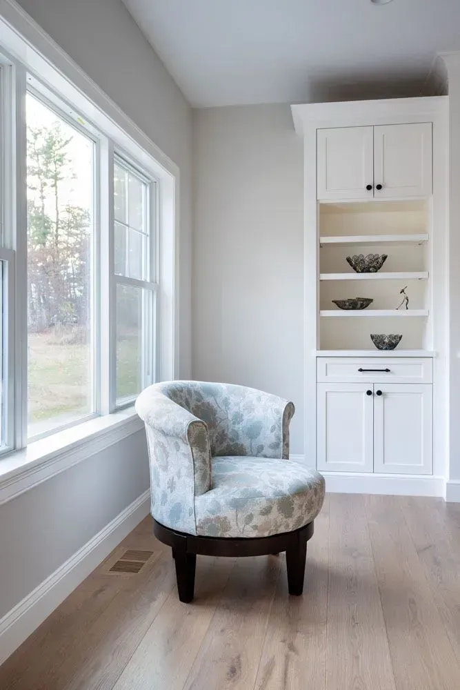 Cozy armchair in front of a window next to a white built-in shelf in a light-filled room with wood floors.