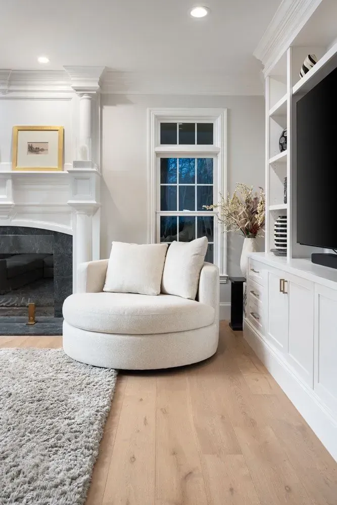 Cozy living room with white swivel chair, built-in shelves, fireplace, hardwood floor, and window with exterior view.