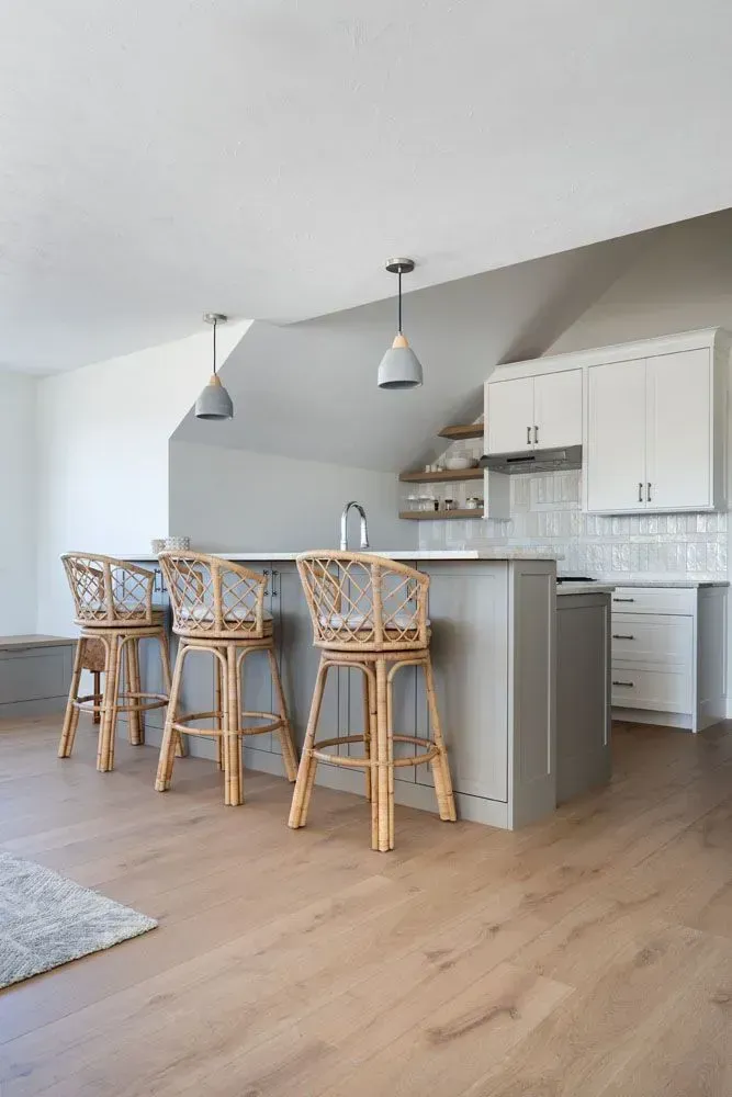Kitchen with gray island, rattan bar stools, light wood floors, and white cabinetry.