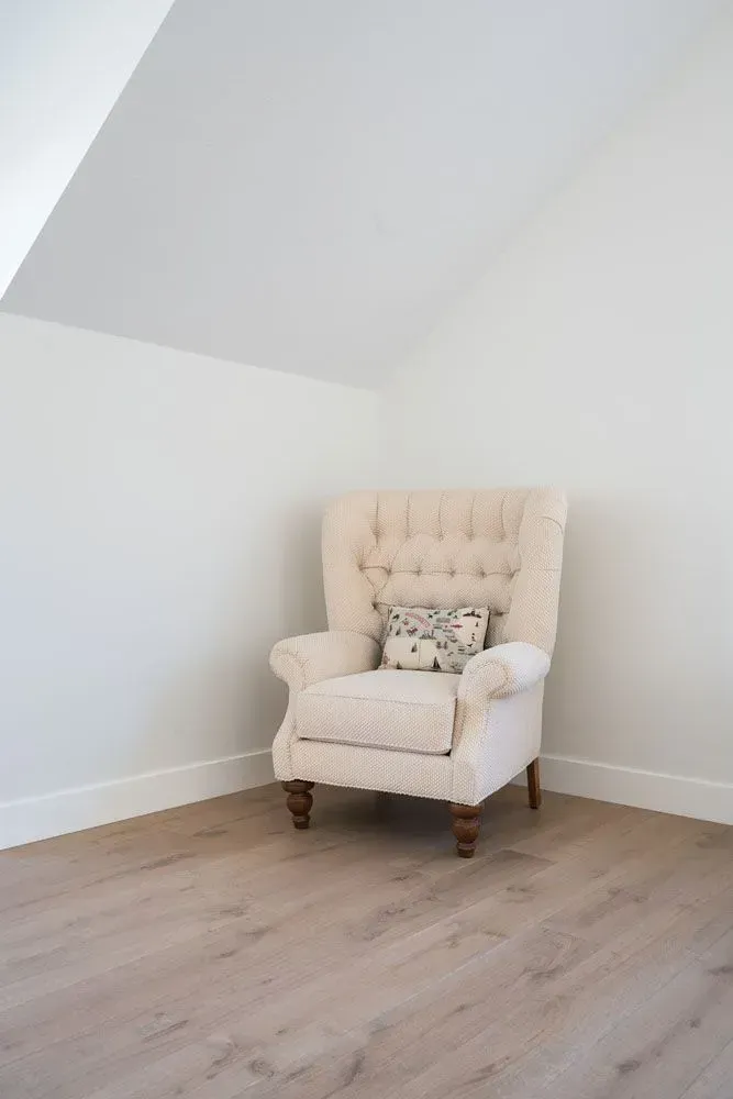 Beige tufted armchair in a corner of a room with white walls and wood-look flooring.