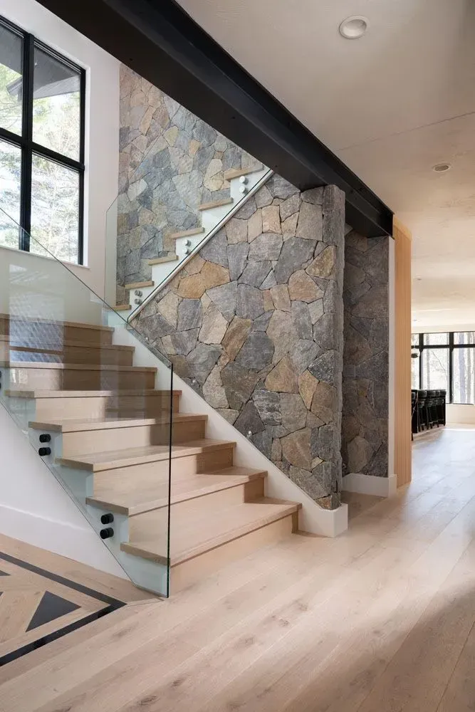Staircase with light wood steps, glass railing, stone wall, and light flooring.