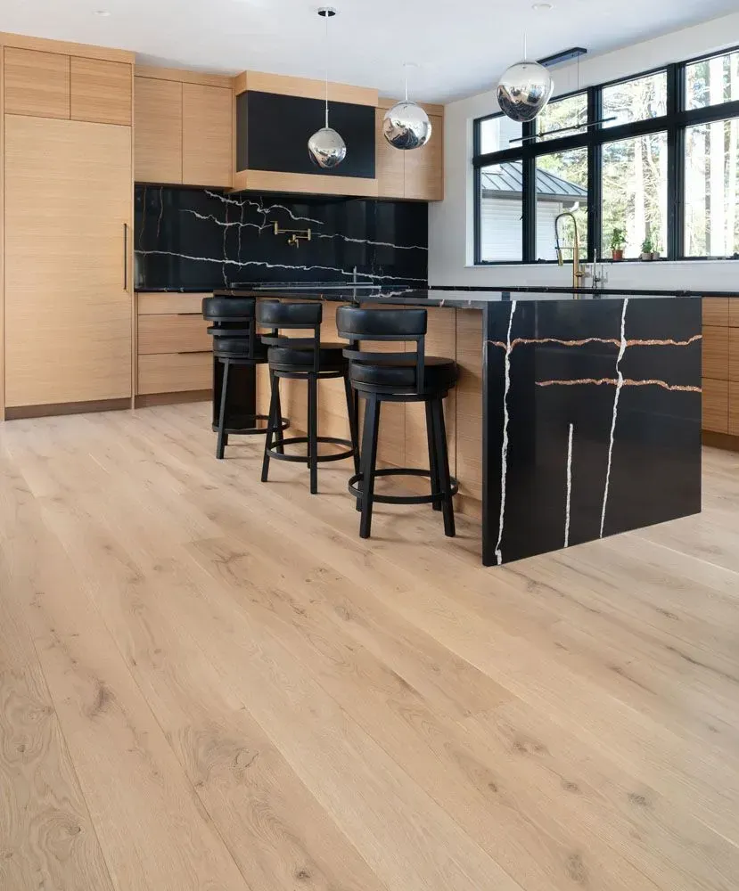 Light wood-floored kitchen with black marble island/backsplash. Wooden cabinets, black stools, silver globe pendants.