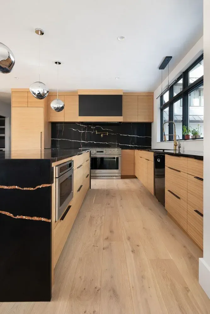 Modern kitchen with light wood cabinets, black countertops, stainless steel appliances, and large windows.