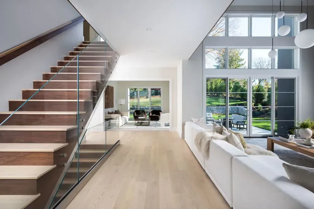 Modern home interior: staircase with glass railing, living room with white sofa, large windows overlooking the yard.
