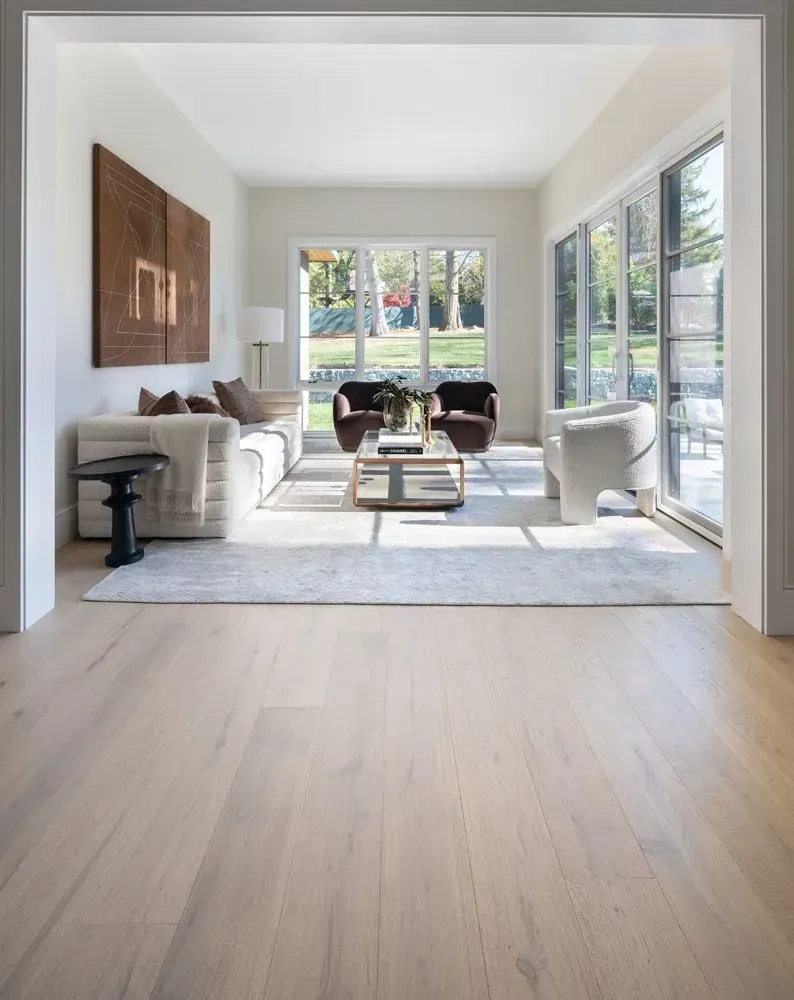 Living room with light wood floors, white walls, and large windows overlooking a yard.
