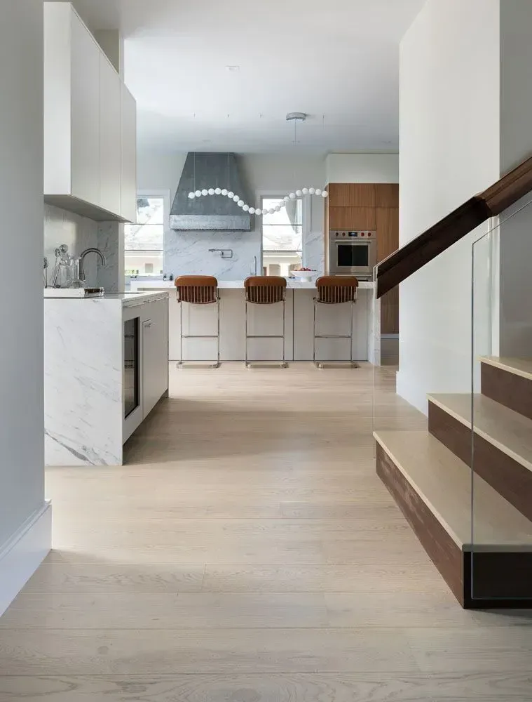 Modern kitchen with white cabinets, marble counters, wooden bar stools, and a staircase.