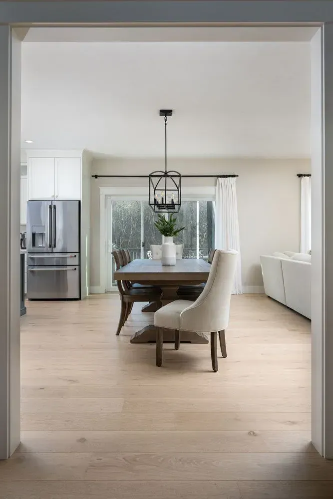 Light-filled dining room with wood floors, table, chandelier, and open doorway to living space.