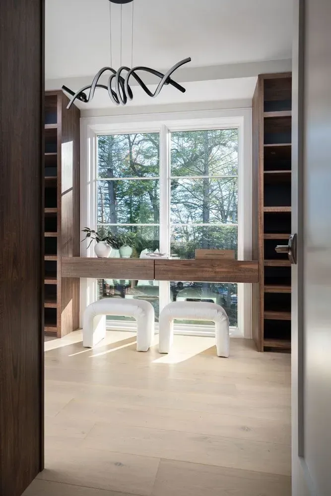 Modern dressing room with a window desk and two white stools, surrounded by built-in wooden shelving.