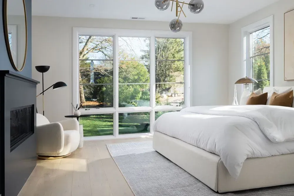 Bedroom with large windows overlooking a green yard, white bed, and modern black fireplace.