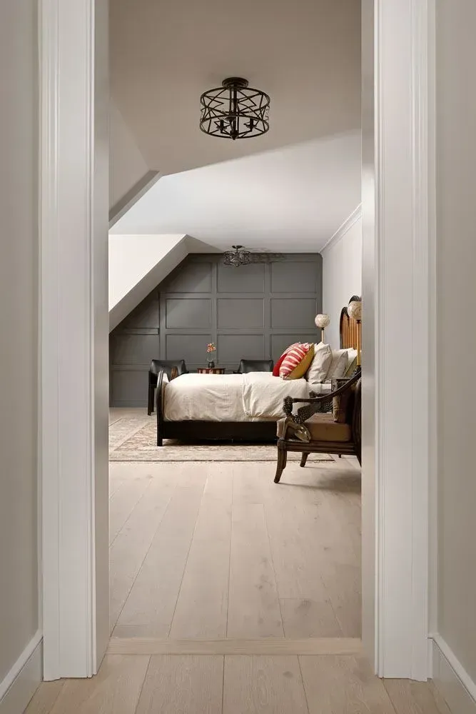 Bedroom viewed from doorway: light wood floors, gray walls, bed with colorful pillows, decorative ceiling lights.