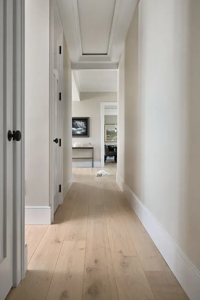 Narrow hallway with light wood floor and white walls, leading to another room.