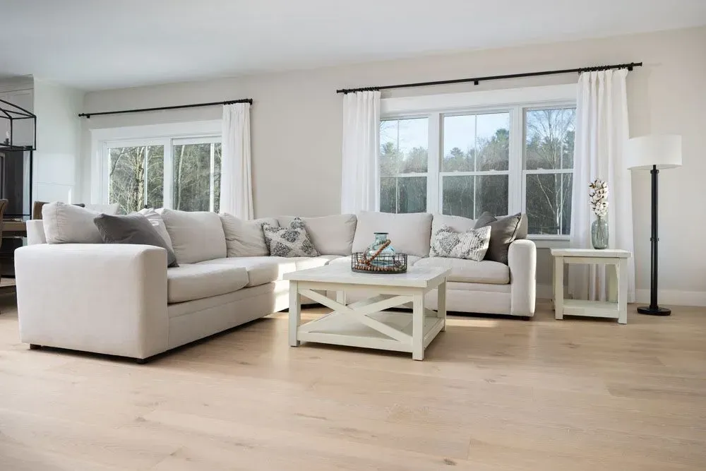 Spacious living room with a light-colored sectional sofa, white walls, and hardwood floors; natural light streams in.