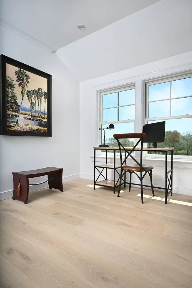 A home office with a desk, art, and a window. Light wood floor, white walls, and a view outside.