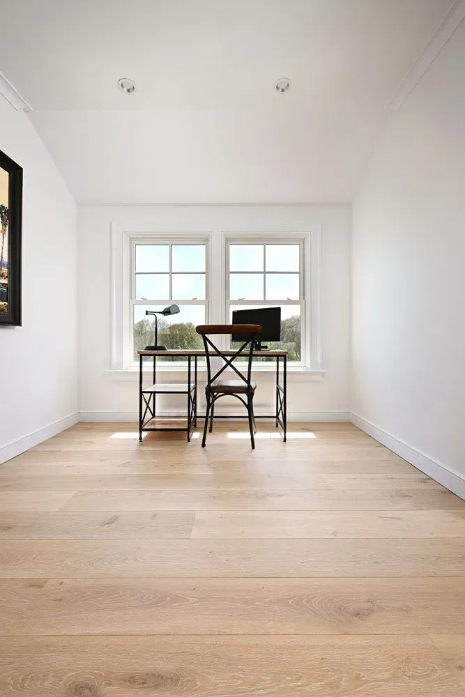 Light-filled home office with a desk, chair, and windows. White walls, light wood floors, and a dark metal desk.
