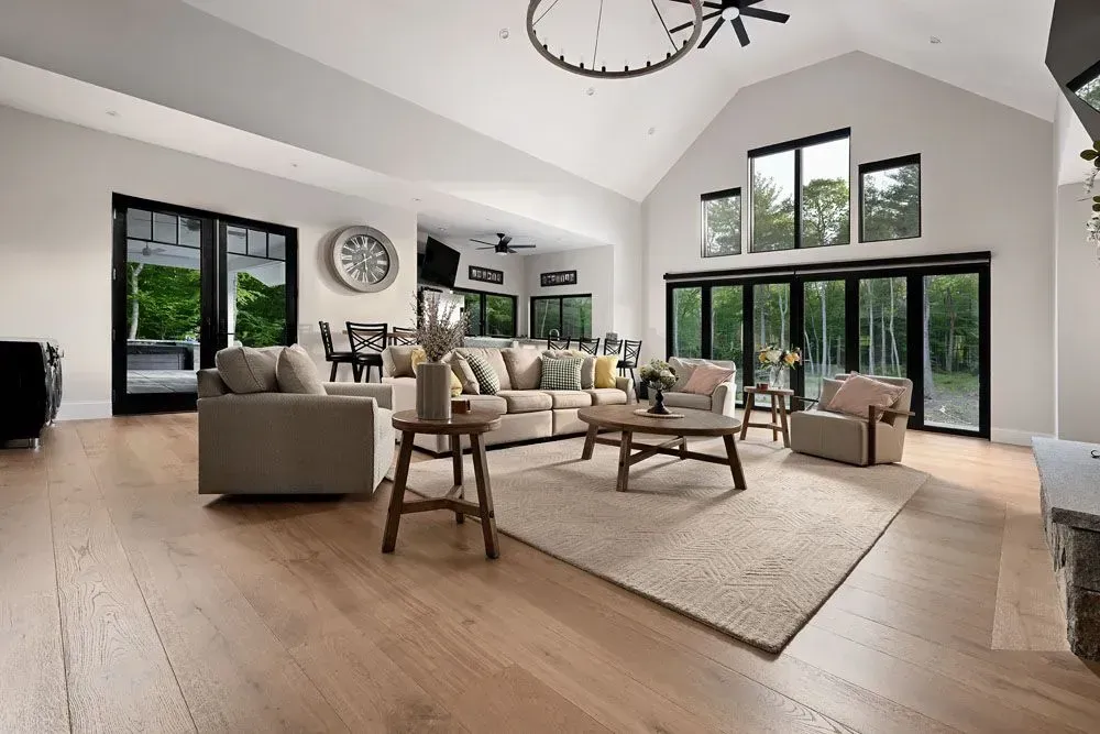 Spacious living room with beige furniture, wooden floors, and large windows overlooking greenery.
