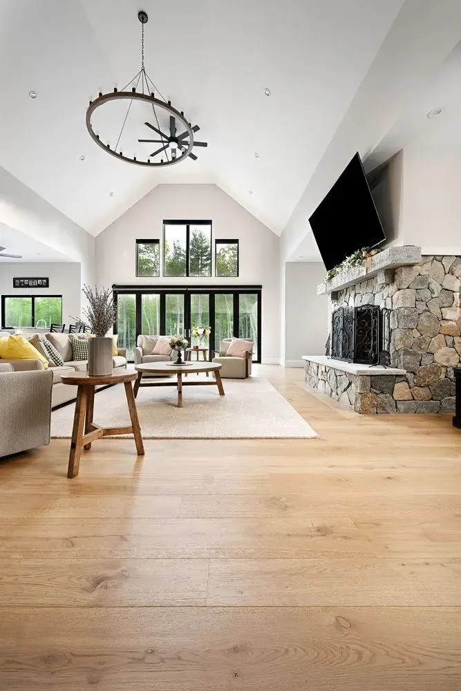 Spacious living room with a stone fireplace, large windows, light wood floors, and a chandelier.