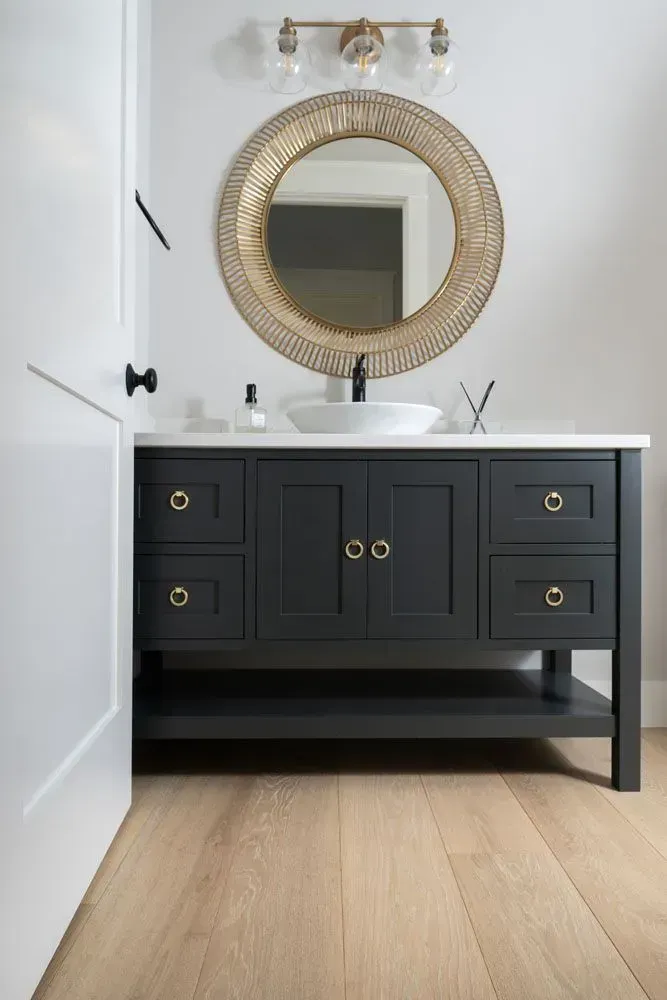 Bathroom vanity with black cabinet, gold hardware, round mirror, and white countertop.