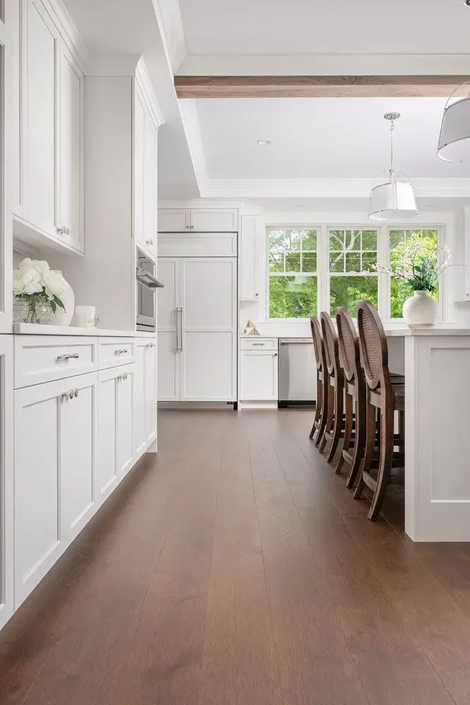 White kitchen with dark wood floor, cabinets, and island seating. Large windows with a view.