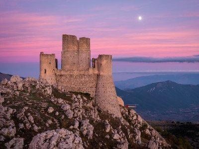 Rovine di un castello sulla cima di una montagna rocciosa al tramonto, con una luna fioca in un cielo rosa e viola.